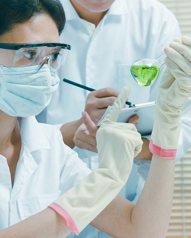 A woman examines a green liquid while wearing glasses, a mask and gloves