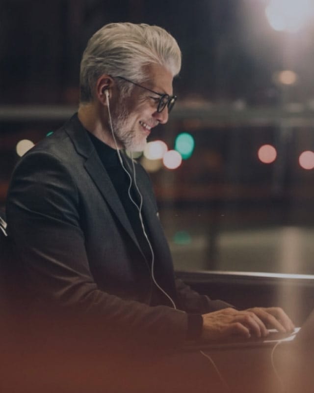 A man smiles with earphones in while looking at a laptop screen